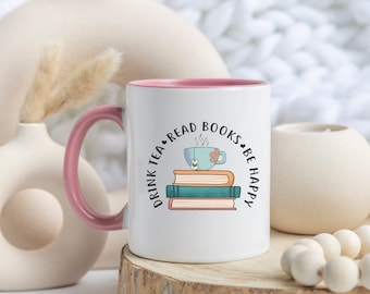 May include: White ceramic mug with a pink rim and handle. The mug has a graphic of a stack of books with a teacup on top. The text around the graphic reads "Drink Tea Read Books Be Happy".