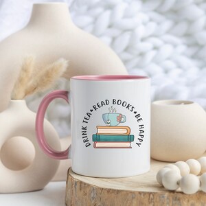 Könnte beinhalten: Weiße Keramiktasse mit rosafarbenem Rand und Henkel. Die Tasse zeigt eine Grafik mit einem Stapel Bücher, auf dem eine Teetasse liegt. Der Text um die Grafik lautet "Drink Tea Read Books Be Happy".