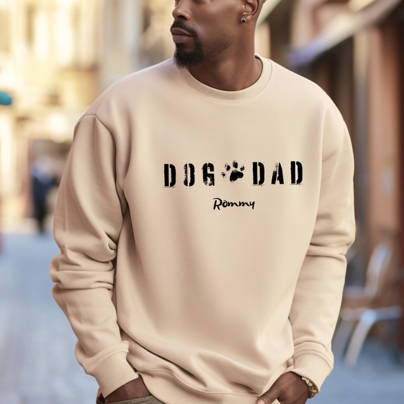 Może przedstawiać: Beżowa bluza z czarnym napisem "DOG DAD", z odciskiem łapy w miejscu "O" w słowie "DOG". Poniżej napisane imię "Rommy" czcionką odręczną. Bluza jest w neutralnym kolorze.