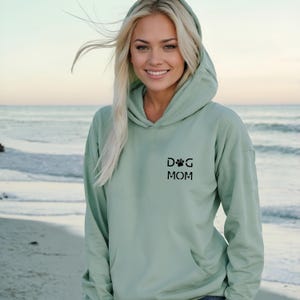 Może przedstawiać: Jasnozielona bluza z kapturem z przednią kieszenią i kapturem. Bluza ma czarny nadruk "DOG MOM". Tło to scena na plaży z oceanem i niebem.