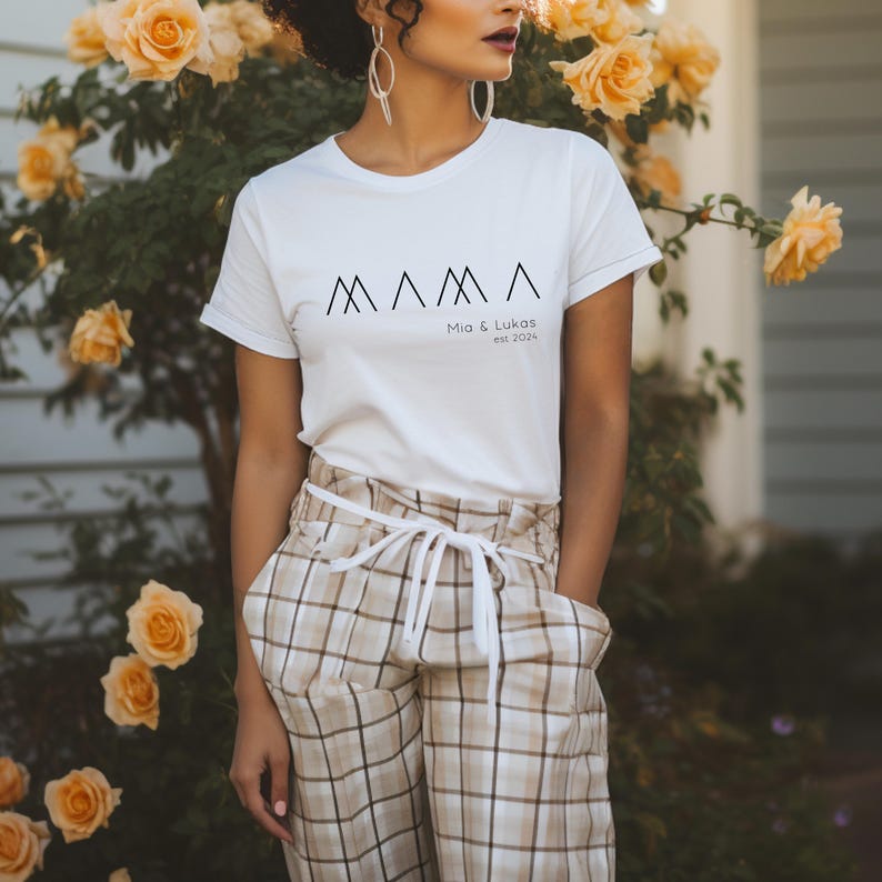 May include: White t-shirt with a black graphic of the word "MAMA" with three triangles. The text "Mia & Lukas est. 2024" is below the graphic.