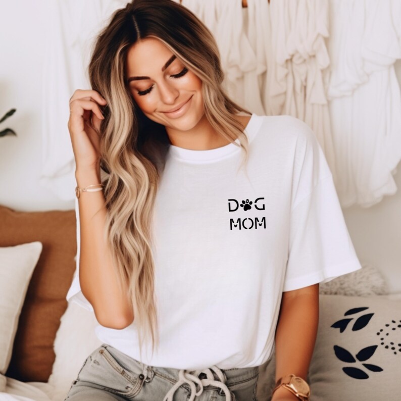 Może przedstawiać: Biała koszulka z napisem "DOG MOM" w kolorze czarnym, z motywem łapy. Koszulka ma swobodny styl, z kr&oacute;tkimi rękawami. Zdjęcie przedstawia koszulkę na osobie.