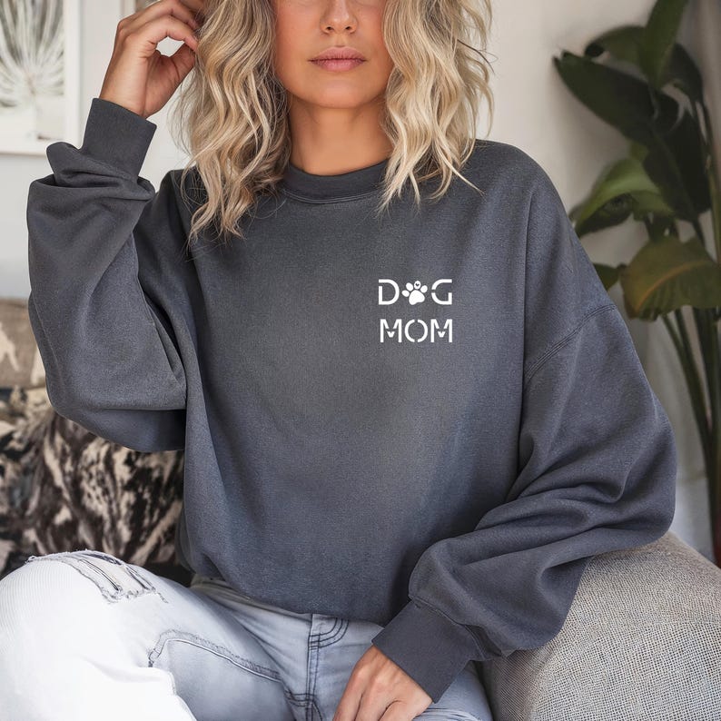 Może przedstawiać: Ciemnoszara bluza z napisem "DOG MOM" w kolorze białym, z motywem łapy. Bluza ma długie rękawy i okrągły dekolt. Odzież wykonana jest z miękkiego materiału.