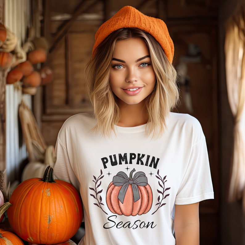May include: A woman wearing a white t-shirt with the text "Pumpkin Season" and a graphic of an orange pumpkin with a gray bow. She is wearing an orange beanie and smiling at the camera.
