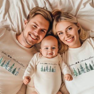 Może przedstawiać: Rodzina trzech osób w pasujących kremowych koszulkach i body. Każdy element garderoby ma akwarelowy wzór drzew iglastych i napis "Frohe Weihnachten" czarnym pismem. Rodzina uśmiecha się i leży na kremowej powierzchni.