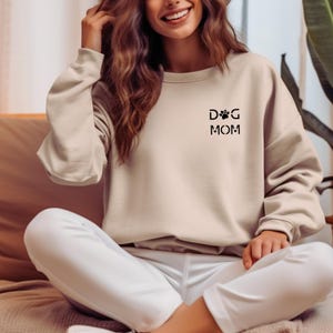 Może przedstawiać: Beżowa bluza z czarnym napisem "DOG MOM". Projekt zawiera odcisk łapy i literę "D" w kształcie łapy. Bluza jest w neutralnym kolorze i ma długie rękawy.