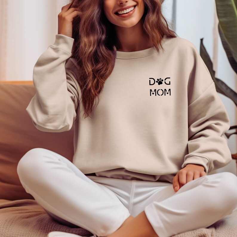 Może przedstawiać: Beżowa bluza z czarnym napisem "DOG MOM". Projekt zawiera odcisk łapy i literę "D" w kształcie łapy. Bluza jest w neutralnym kolorze i ma długie rękawy.