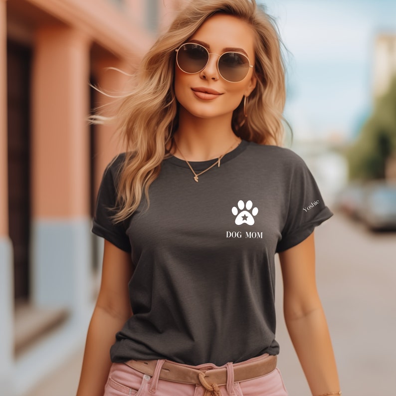 May include: A dark gray t-shirt with a white paw print and the text "DOG MOM" on the front. The t-shirt is being worn by a person with long blonde hair and sunglasses.