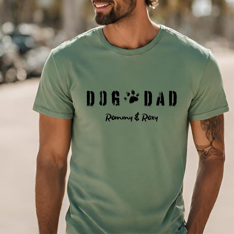 Może przedstawiać: Szałwiowa zielona koszulka z napisem "DOG DAD" w stylu distressed, z motywem odcisku łapy. Poniżej tekstu znajduje się imię "Rommy & Roxy" w czcionce skryptowej. Koszulka jest w stylu casual.