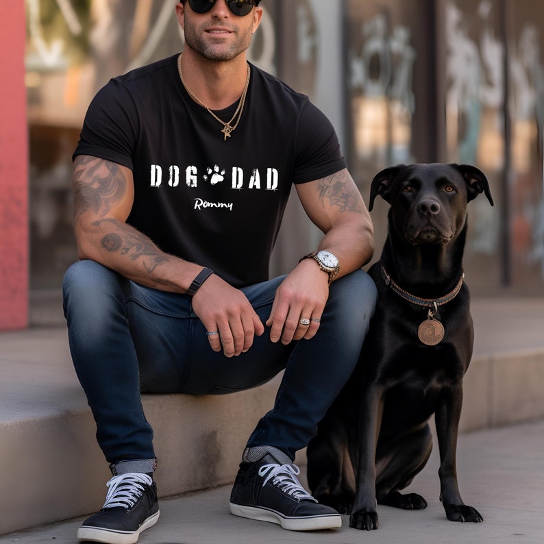 Może przedstawiać: Czarny t-shirt z napisem "DOG DAD" i wzorem łapy. Imię "Rommy" jest wydrukowane pod tekstem. Czarny pies z brązową obrożą siedzi obok osoby noszącej koszulkę.