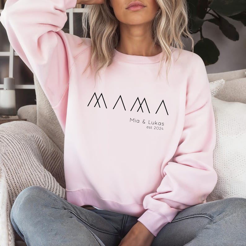 May include: Pink sweatshirt with the word "MAMA" in black stylized letters, with the text "Mia & Lukas est 2024" below.