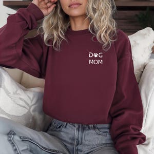 Może przedstawiać: Bordowa bluza z okrągłym dekoltem z białym napisem "DOG MOM". Projekt zawiera odcisk łapy i kość dla psa. Bluza wykonana jest z miękkiego materiału i nadaje się do noszenia na co dzień.