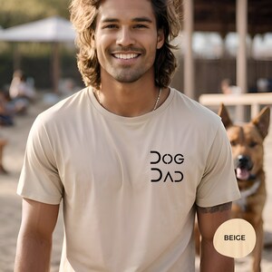 Pu&ograve; includere: Una maglietta beige con una grafica nera che dice "DOG DAD" in un font stilizzato. La maglietta &egrave; indossata da una persona con i capelli lunghi castani. La parola "BEIGE" &egrave; scritta su un cerchio marrone nell'angolo in basso a destra dell'immagine.