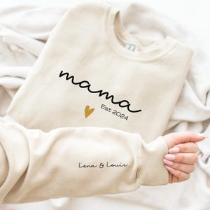 Op de afbeelding: Een beige sweatshirt met de tekst "mama Est 2024" en een klein gouden hartje. De mouw heeft de tekst "Lena & Louis" in zwart.
