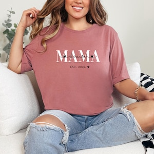 Może przedstawiać: Kobieta w malinowym t-shircie z napisem "MAMA" i "doula" w białym kolorze. Na koszulce znajduje się r&oacute;wnież napis "EST. 2024" z małym symbolem serca.