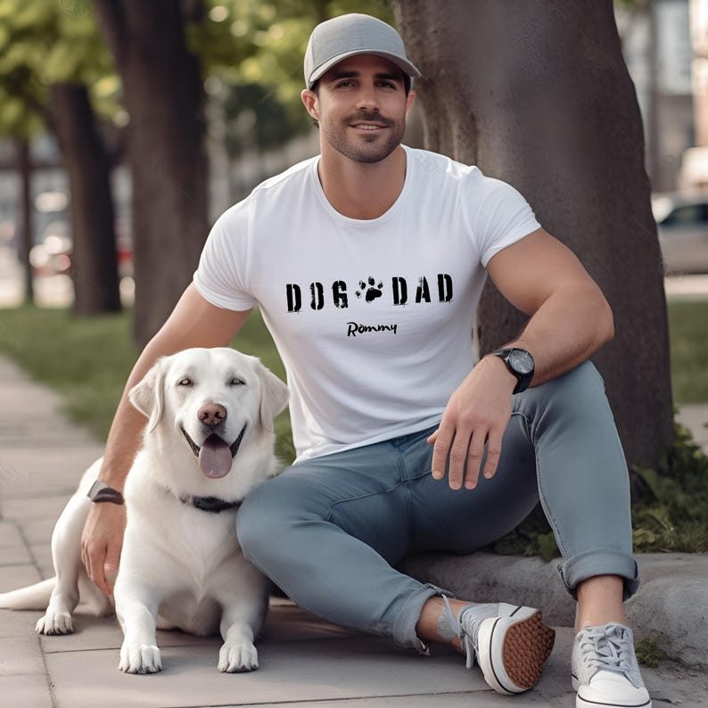 Może przedstawiać: Biała koszulka z napisem "DOG DAD" i wzorem łapy. Imię "Rommy" jest wydrukowane poniżej. Koszulka jest noszona przez osobę siedzącą obok białego psa rasy Labrador Retriever.