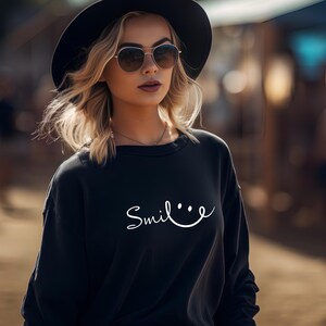 Pode incluir: Um moletom preto com a palavra "Smile" em escrita branca. A pessoa na imagem está usando óculos de sol e um chapéu preto. O fundo está desfocado, sugerindo um ambiente ao ar livre.