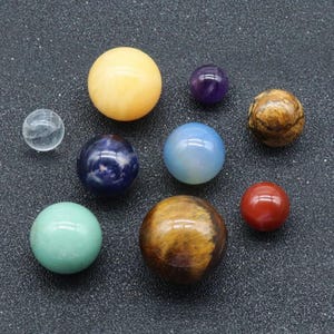 May include: A collection of ten smooth, polished gemstone spheres in various colors, including clear, yellow, blue, green, brown, red, and purple. The spheres are arranged on a dark gray surface.