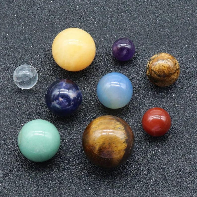 May include: A collection of ten smooth, polished gemstone spheres in various colors, including clear, yellow, blue, green, brown, red, and purple. The spheres are arranged on a dark gray surface.