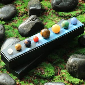 May include: A set of eight gemstone spheres representing the planets in our solar system. The spheres are arranged on a black box with the names of the planets printed on it. The planets are Mercury, Venus, Earth, Mars, Jupiter, Saturn, Uranus, and Neptune.