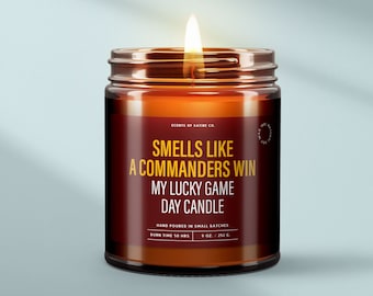 May include: A lit candle in a glass jar with a brown label. The label reads "Smells Like a Commanders Win My Lucky Game Day Candle." The candle is 9 oz. and has a burn time of 50 hours. The jar is on a light blue background.
