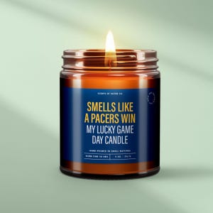 May include: A brown glass candle with a blue label that reads "Smells like a Pacers win, My Lucky Game Day Candle". The candle is lit and has a white wick. The label also says "Hand poured in small batches, Burn time 50 hrs, 9 oz / 252 g".