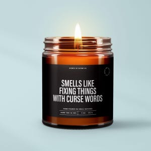 May include: A lit candle in an amber glass jar with a black label. The label reads: SCENTS OF SATIRE CO. SMELLS LIKE FIXING THINGS WITH CURSE WORDS. HAND POURED IN SMALL BATCHES. BURN TIME 50 HRS. 9 OZ. / 252 G. WAX 100% NATURAL SOY.