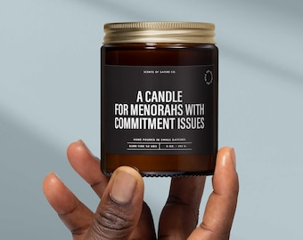 May include: A brown glass jar candle with a gold lid, held in a hand. The black label reads "A CANDLE FOR MENORAHS WITH COMMITMENT ISSUES." The candle is hand-poured in small batches and has a burn time of 50 hours. The candle is 8 oz / 212 g.