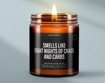 May include: A lit candle in a glass jar with a black label. The label reads "Smells Like Eight Nights of Chaos and Carbs." The candle is hand-poured and has a burn time of 50 hours. The jar contains 9 oz / 252 g of wax.