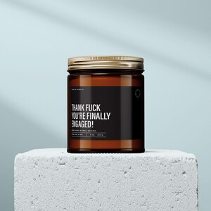 May include: A brown glass jar candle with a gold lid. The black label reads "THANK FUCK YOU'RE FINALLY ENGAGED!" in white text. The candle is on a white block against a light blue background. The candle is 8 oz.