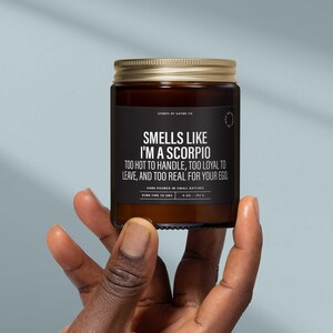 May include: A brown glass jar candle with a gold lid, held in a hand. The black label reads "Smells Like I'm a Scorpio" with additional text. The candle is 8 oz. and has a 50-hour burn time.