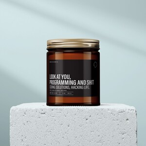May include: A brown glass candle jar with a gold lid. The label reads "LOOK AT YOU, PROGRAMMING AND SHIT CODING SOLUTIONS, HACKING LIFE." The candle is on a white surface.