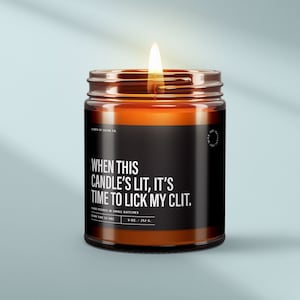 May include: A brown glass candle with a black label that reads "WHEN THIS CANDLE'S LIT, IT'S TIME TO LICK MY CLIT." The candle is lit and the flame is visible. The label also includes the text "HAND POURED IN SMALL BATCHES" and "9 OZ. / 252 G."