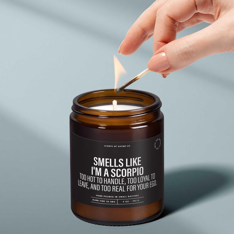 May include: A lit candle in a brown glass jar with a black label. The label reads "Smells Like I'm a Scorpio." A hand holds a lit match above the candle. The candle is hand-poured and has a burn time of 50 hours.