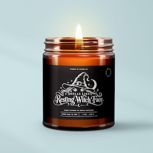 May include: A lit candle in an amber glass jar with a black label. The label features a white graphic of a witch hat and the text "Scents of Satire Co. Smells Like Resting Witch Face. Hand Poured in Small Batches. Burn Time 50 Hrs. 9 Oz. / 252 G."