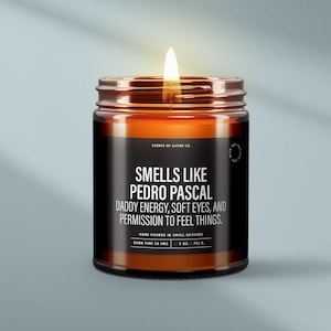 May include: A lit candle in a glass jar with a black label. The label reads "Smells Like Pedro Pascal, Daddy Energy, Soft Eyes, and Permission to Feel Things." The candle is hand-poured and has a burn time of 50 hours. It weighs 9 oz / 252 g.