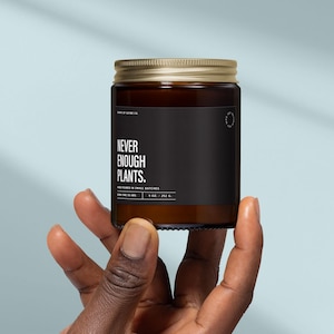May include: A brown glass candle jar with a gold lid and a black label. The label reads "NEVER ENOUGH PLANTS." The candle is 9 oz. / 252 g. The jar is held in a hand against a light blue background.