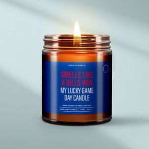 May include: A brown glass candle jar with a blue label that reads "Smells like a Bills win My Lucky Game Day Candle". The label also includes the text "Scents of Satire Co.", "Hand poured in small batches", "Burn time 50 hrs", and "9 oz./252 g".