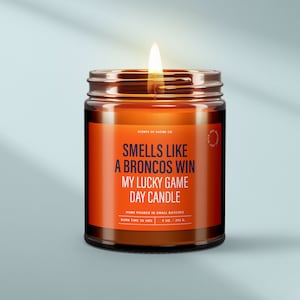 May include: A brown glass candle jar with a white label that reads "Smells Like A Broncos Win My Lucky Game Day Candle". The label also includes the text "Scents of Satire Co.", "Hand Poured in Small Batches", "Burn Time 50 Hrs", and "9 oz / 252 g".