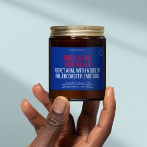 May include: A brown glass candle jar with a gold lid. The label is blue with white text that reads "SCENTS OF SATIRE CO. SMELLS LIKE JOSH ALLEN ROCKET ARM, WITH A SIDE OF ROLLERCOASTER EMOTIONS HAND POURED IN SMALL BATCHES BURN TIME 50 HRS 4 OZ / 252 G."