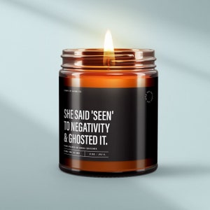 May include: A brown glass candle with a black label that reads "She said 'seen' to negativity & ghosted it." The candle is lit and the flame is visible. The label also includes the text "Hand poured in small batches" and "9 oz. / 252 g."