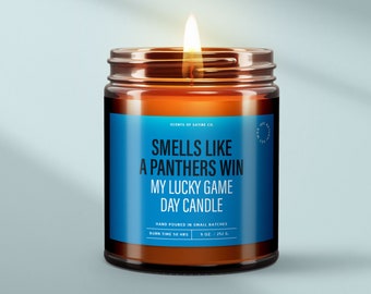 May include: A brown glass candle jar with a blue label that reads "Smells like a Panthers win my lucky game day candle". The label also includes the text "Scents of Satire Co.", "Hand poured in small batches", "Burn time 50 hrs", and "9 oz./252 g".