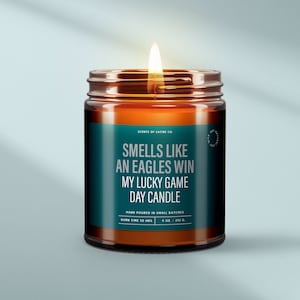 May include: A brown glass candle with a teal label that reads "Smells like an Eagles win my lucky game day candle". The candle is hand-poured in small batches and has a burn time of 50 hours. It is 9 ounces or 252 grams.