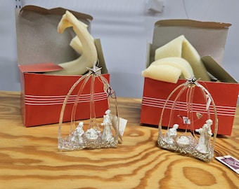 May include: Two glass nativity scene ornaments featuring white figures of Mary, Joseph, and the infant Jesus under a clear arched frame topped with a silver star. Each ornament is accompanied by its own red cardboard box with foam padding.