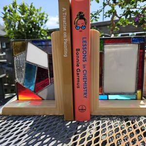 May include: Wooden bookends with stained glass panels in a geometric design. The bookends hold two books: "A Treatise on Painting" and "Lessons in Chemistry" by Bonnie Garmus. The bookends are on a metal surface.