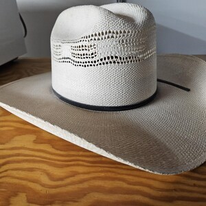 May include: A white straw cowboy hat featuring a cattleman crease, a wide brim, and a ventilated crown section. The hat includes a thin black band encircling the base of the crown, providing a contrast to the light-colored woven straw material.