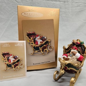 May include: A gold-colored sleigh ornament with Santa Claus inside. Santa is wearing a red suit and a black hat. He is holding a telescope and looking out of the sleigh. The sleigh is decorated with green and red accents. The ornament is labeled "Plotting the Course" and is a Hallmark Club Exclusive.