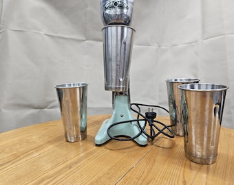 May include: Vintage chrome milkshake maker with a light blue base and three stainless steel cups. The mixer has a tall, cylindrical design with a motor housing at the top. The cups are tapered and have a shiny finish.