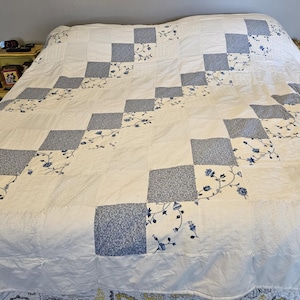 May include: A large bed quilt featuring a patchwork design of solid white squares alternating with blue and white floral patterned squares. The quilt is arranged in a diagonal grid pattern, covering the entire surface of a bed with a white border.