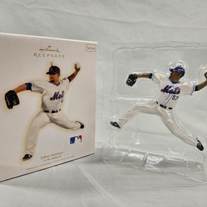 May include: A Hallmark Keepsake ornament featuring Johan Santana, a baseball pitcher for the New York Mets. The ornament is in a clear plastic packaging with a white box behind it. The box has the Hallmark Keepsake logo and the text "Johan Santana At the Ballpark".